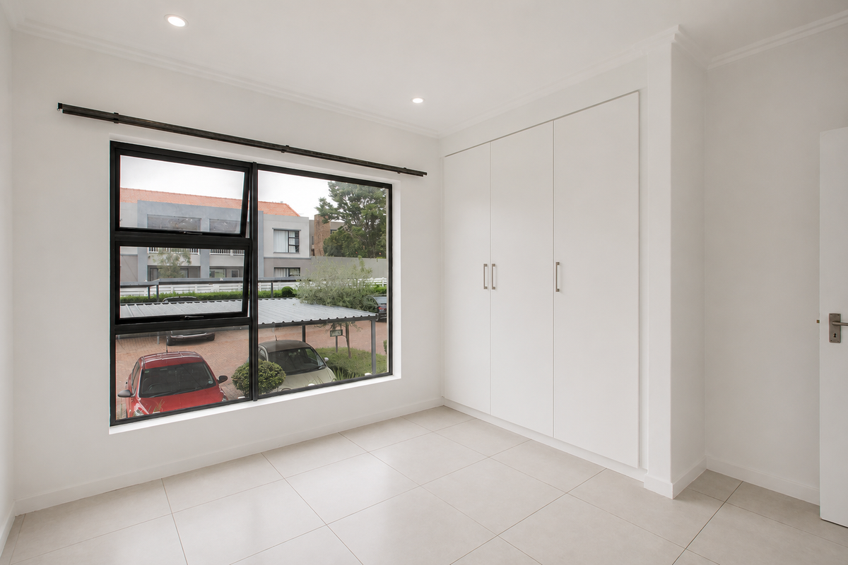 A bright, empty bedroom featuring neutral tiled flooring, built-in cupboards, and a large window with a view of the complex parking area.