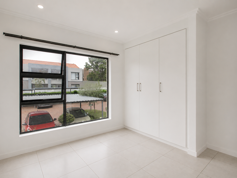 A bright, empty bedroom featuring neutral tiled flooring, built-in cupboards, and a large window with a view of the complex parking area.
