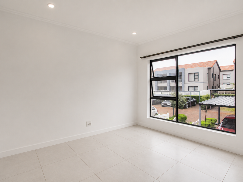 A bright, empty bedroom featuring neutral tiled flooring, recessed downlights, and a large window with black aluminium frames overlooking the complex parking area.