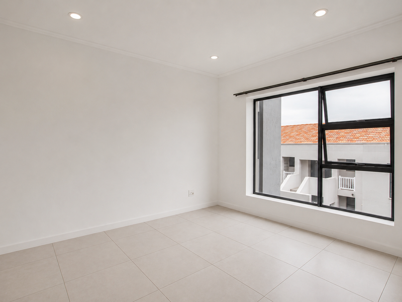 A bright, empty bedroom featuring neutral tiled flooring, recessed downlighting, and a large window with a black aluminium frame.