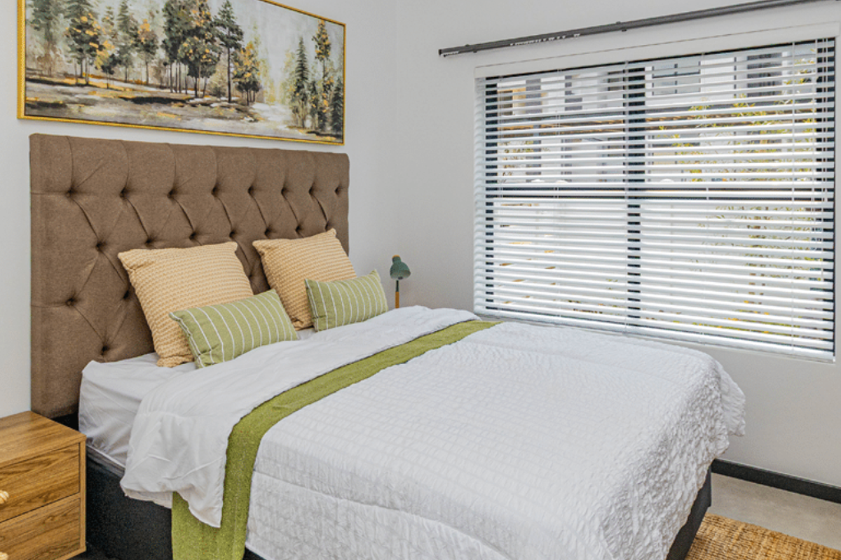 A well-lit bedroom featuring neutral walls, a large window with fitted venetian blinds, and a modern curtain rod.