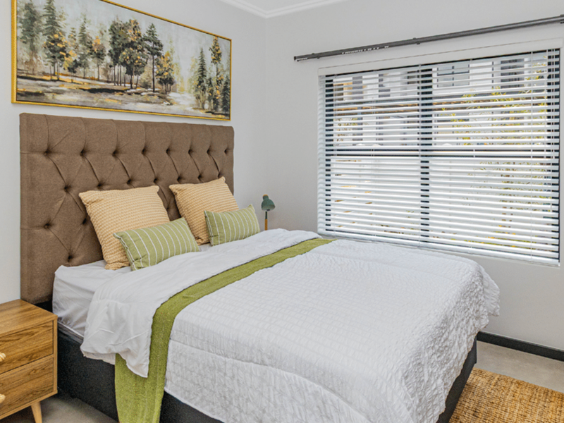 A well-lit bedroom featuring neutral walls, a large window with fitted venetian blinds, and a modern curtain rod.