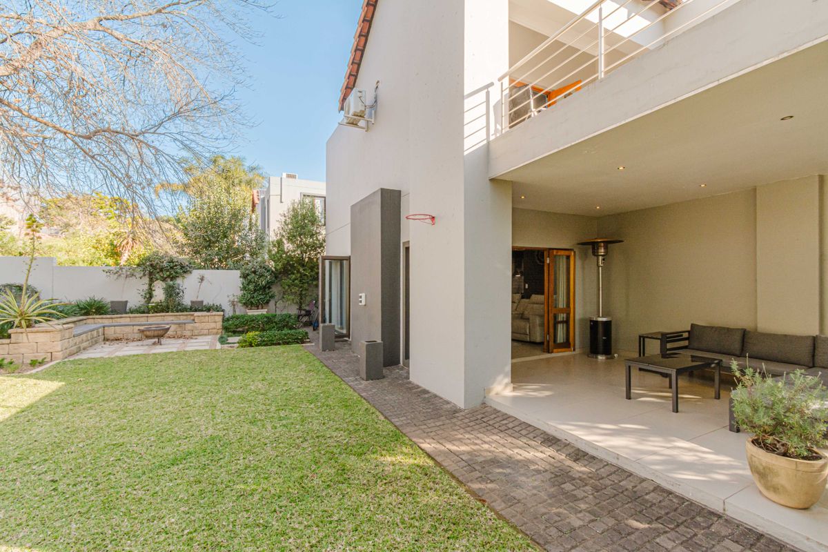Rear view of the property featuring a covered patio, balcony, paved walkway, and a well-maintained garden with a retaining wall.