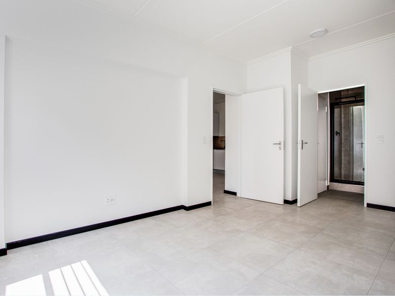 A bright, empty bedroom featuring neutral tiled flooring, clean white walls, and direct access to an adjoining bathroom and kitchen area.