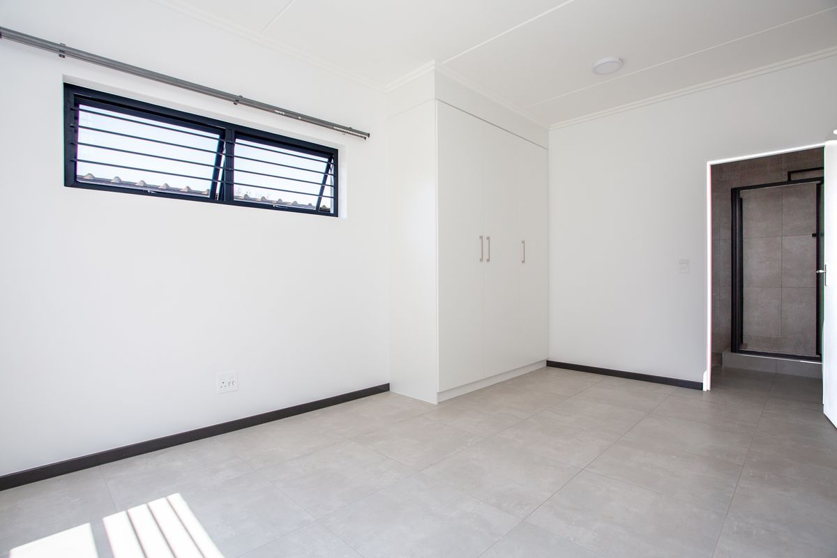 A bright, empty bedroom featuring neutral tiled flooring, a built-in wardrobe, and a louvre window with security bars, leading to an en-suite bathroom.
