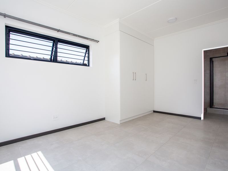 A bright, empty bedroom featuring neutral tiled flooring, a built-in wardrobe, and a louvre window with security bars, leading to an en-suite bathroom.