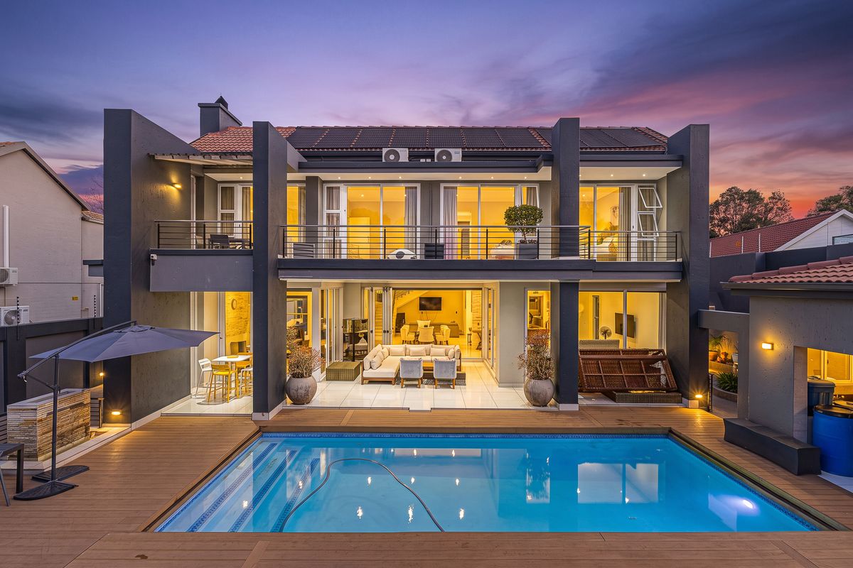 A modern double-story rear elevation featuring a swimming pool, timber decking, a spacious covered patio, and an upper-level balcony with glass sliding doors.
