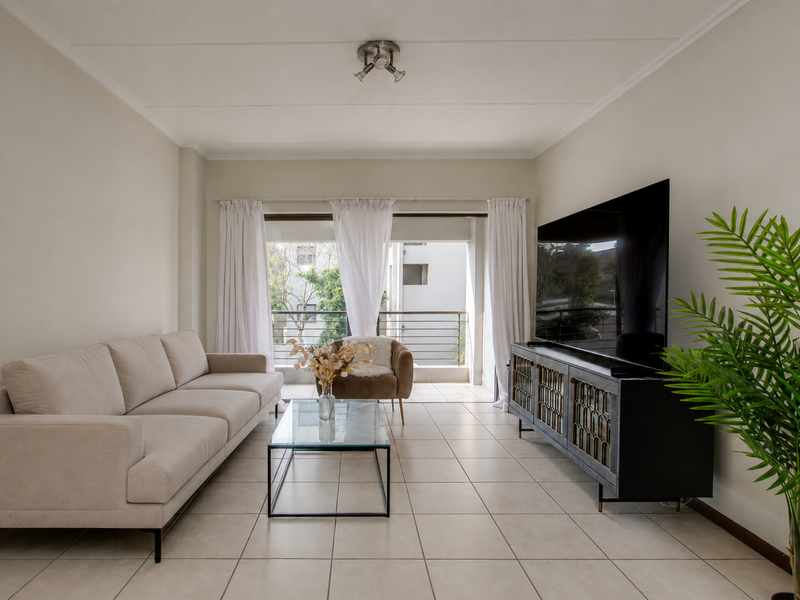 A bright and well-maintained living room featuring neutral tiled flooring and direct access to a private balcony through a large sliding glass door.