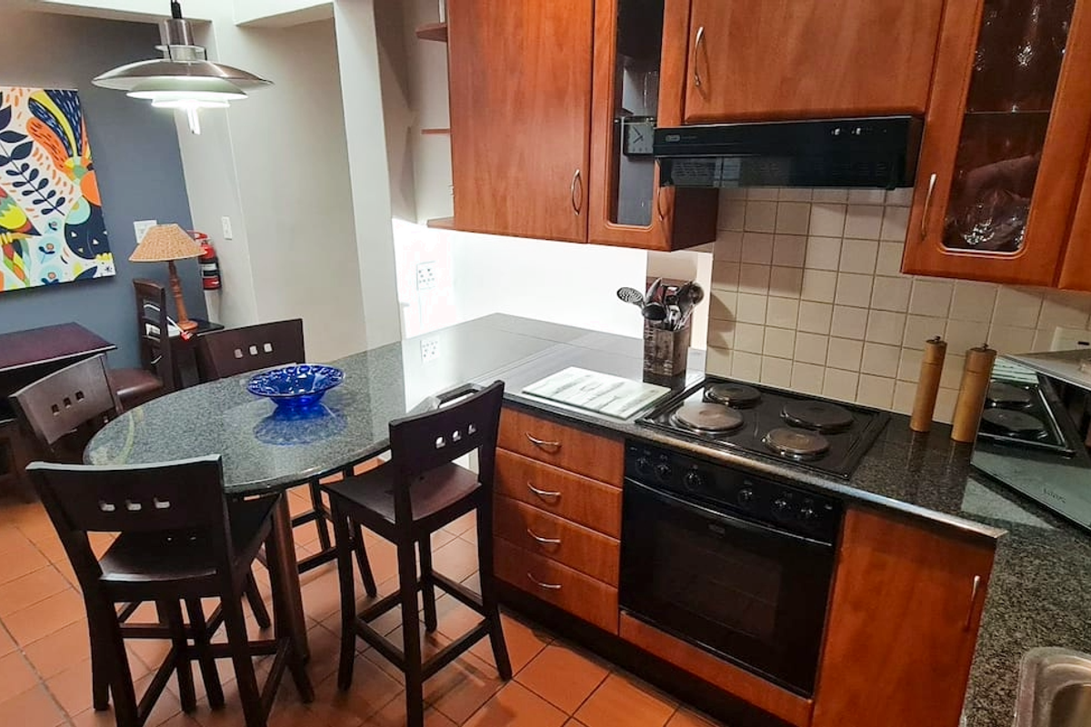 A functional kitchen featuring wooden cabinetry, granite countertops, an integrated electric hob and oven, and tiled flooring with an adjacent dining area.