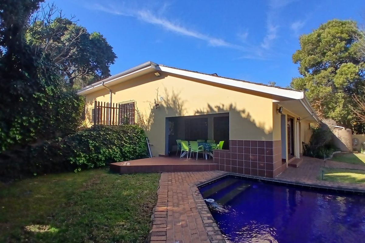 Rear exterior view of the guest house featuring a swimming pool, brick paved walkway, and a covered patio area set within a garden.