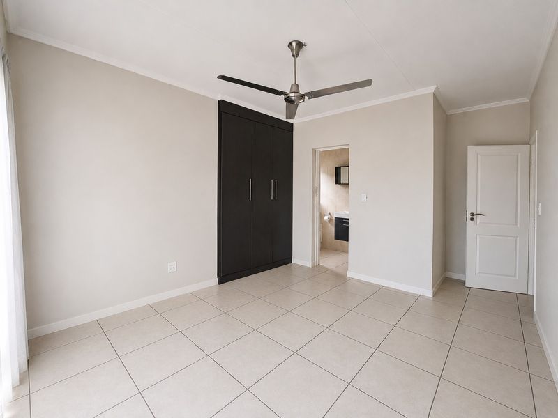 A bright, empty bedroom featuring neutral tiled flooring, built-in dark cabinetry, a modern ceiling fan, and direct access to an en-suite bathroom.