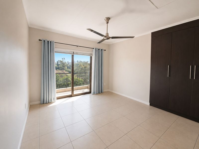 A bright, empty bedroom featuring neutral tiled flooring, a dark built-in wardrobe, a ceiling fan, and sliding door access to a private balcony.