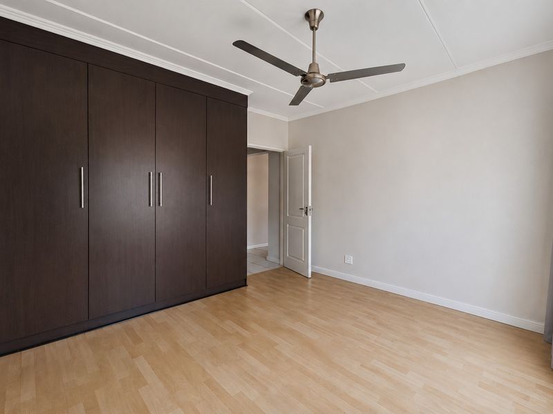 A bright, empty bedroom featuring dark wood built-in cupboards, a modern ceiling fan, and light-toned laminate flooring.