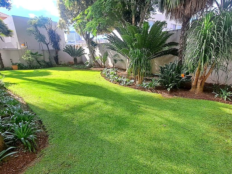 A well-maintained private garden featuring a lush green lawn, established trees, and landscaped garden beds enclosed by a perimeter boundary wall.