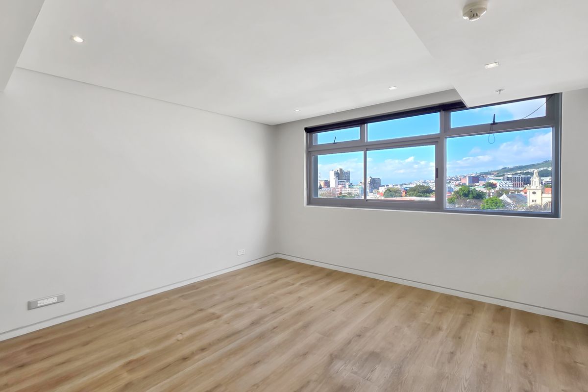 A bright, empty bedroom featuring modern laminate flooring, recessed ceiling lights, and large windows offering expansive city views.