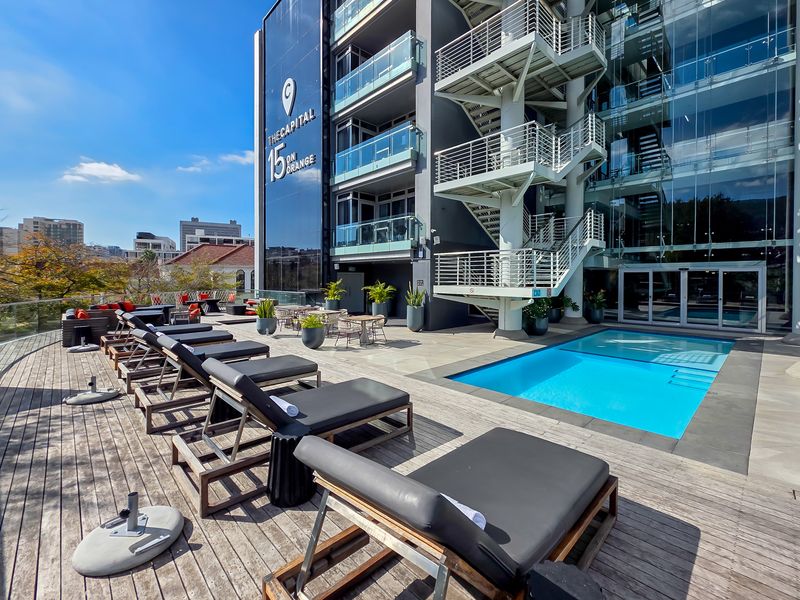 A modern penthouse pool deck featuring timber decking, glass balustrades, and an external staircase, set against a contemporary glass-facade building.