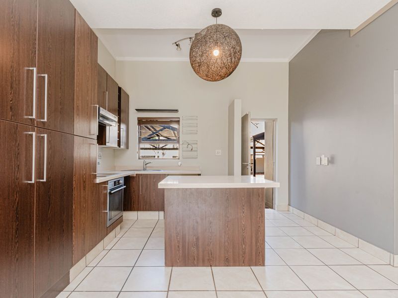 A modern kitchen featuring dark wood cabinetry, a central island with stone countertops, built-in oven, extractor fan, and neutral tiled flooring.