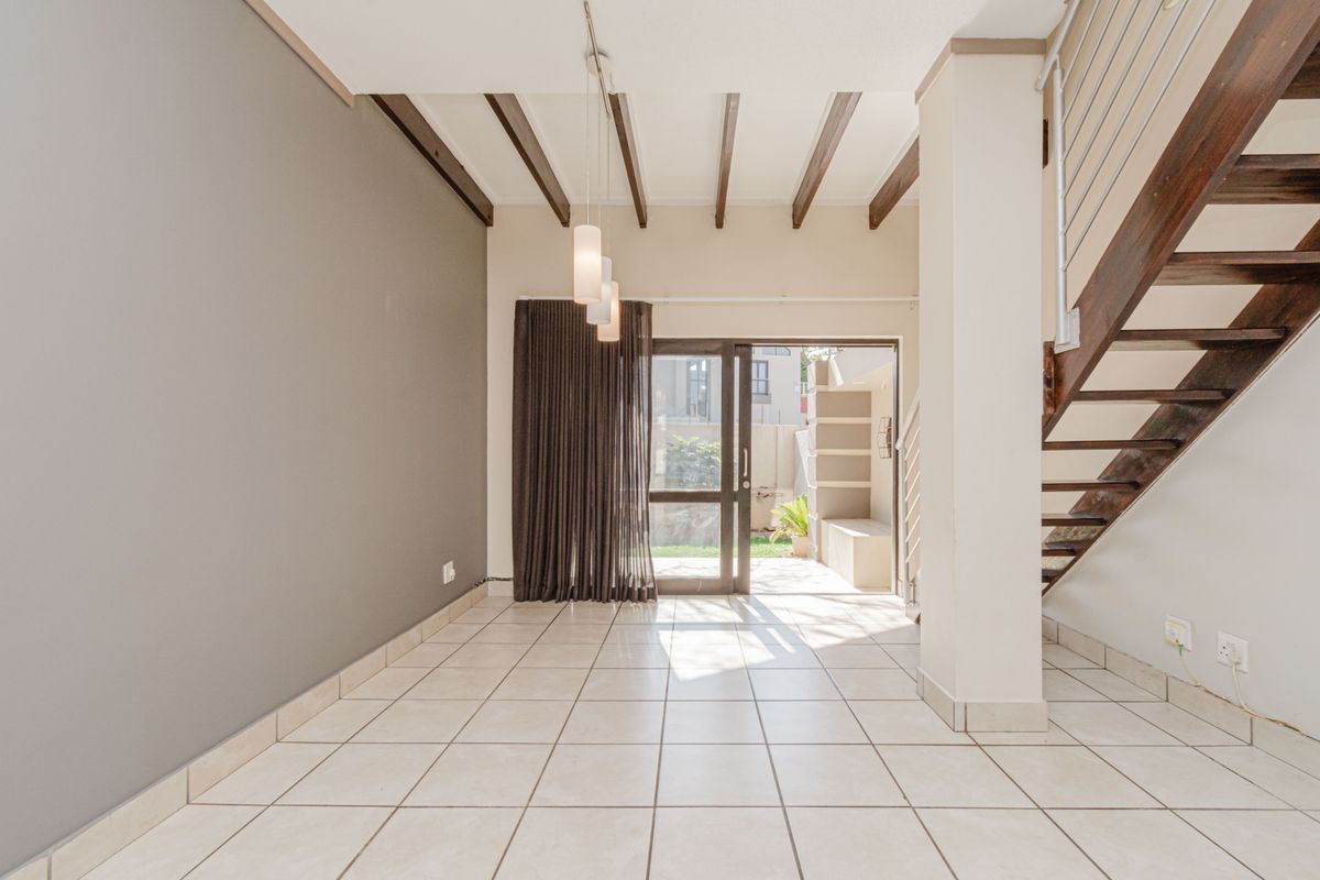 An open-plan living area featuring tiled floors, exposed timber ceiling beams, and a staircase, with direct access to an outdoor patio area.
