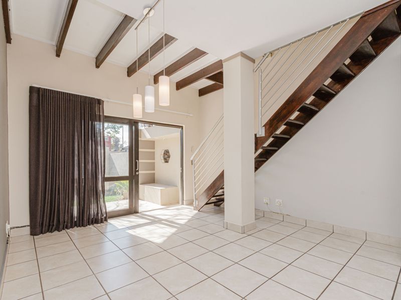 Bright open-plan living area featuring exposed timber ceiling beams, tiled flooring, and a feature staircase leading to the upper level with garden access.