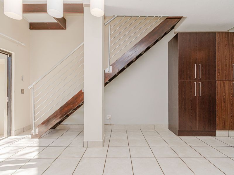 An open-plan living area featuring a modern staircase with metal balustrades, tiled flooring, exposed ceiling beams, and built-in dark wood cabinetry.