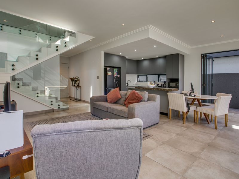 Modern open-plan living area featuring a sleek glass staircase, contemporary kitchen with dark cabinetry, and seamless access to the outdoor patio through bi-fold doors.