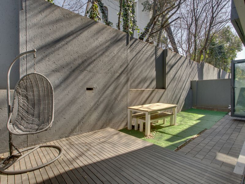 A private outdoor patio area featuring timber decking, paved sections, artificial grass, and a high concrete boundary wall with integrated electric fencing.