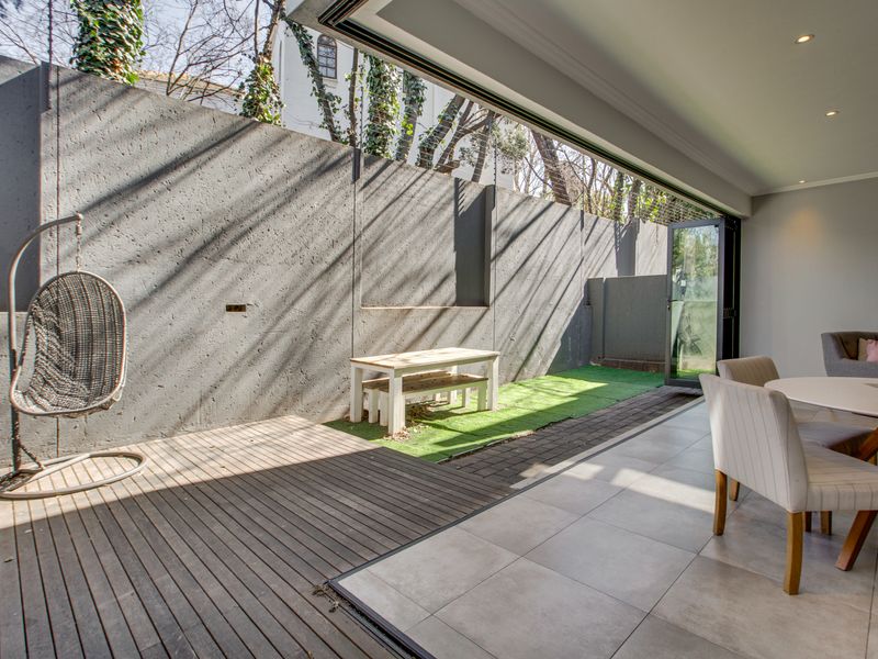 A modern indoor-outdoor living space featuring timber decking, tiled flooring, and seamless stacking glass doors opening to a private courtyard with a concrete wall.