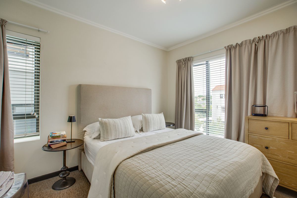 A well-lit bedroom featuring neutral walls, carpeted flooring, and dual windows with fitted blinds and curtains, offering a comfortable and functional living space.