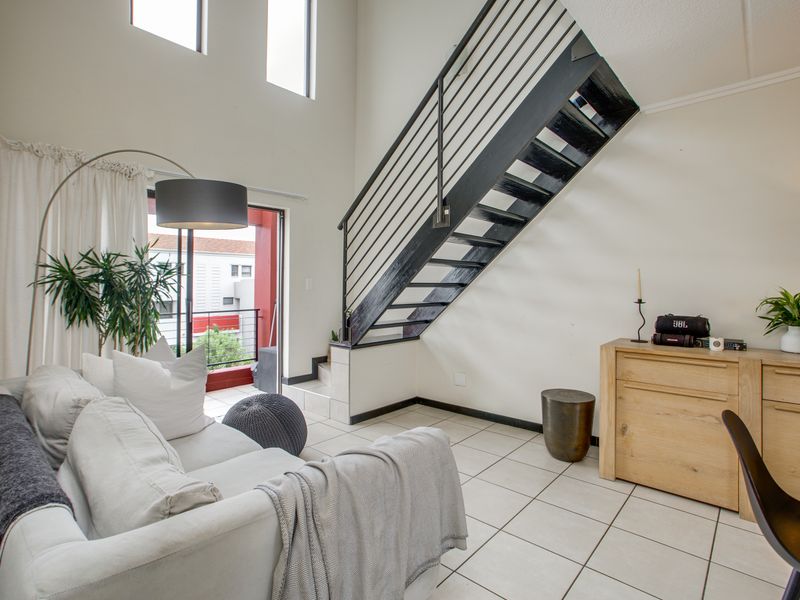 A bright, double-volume living area featuring tiled floors, a modern metal staircase, and direct access to a private balcony through sliding glass doors.