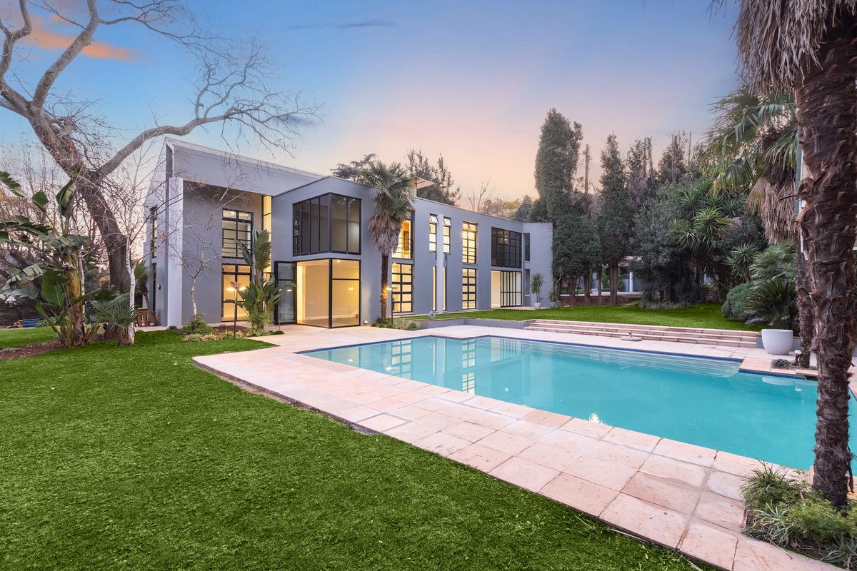 A modern two-story residence featuring a large swimming pool, stone patio, and expansive glass windows overlooking a well-maintained garden.
