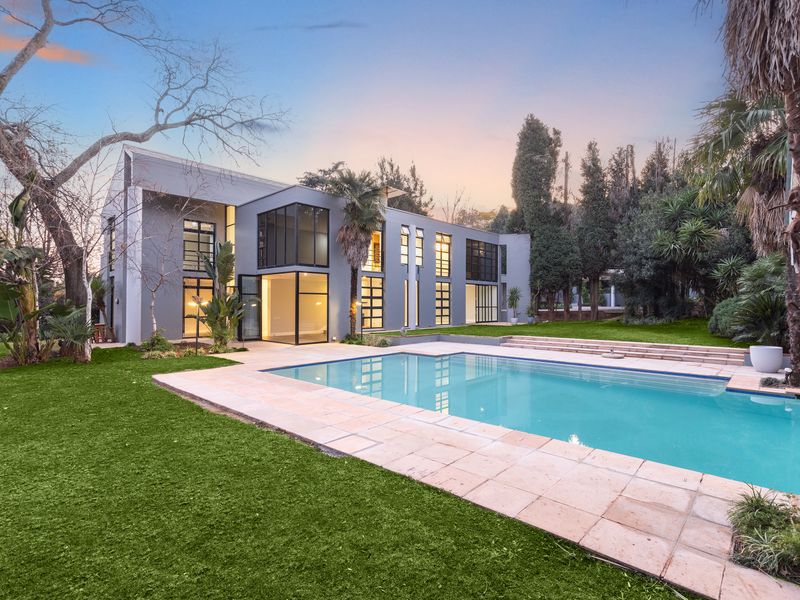 A modern two-story residence featuring a large swimming pool, stone patio, and expansive glass windows overlooking a well-maintained garden.
