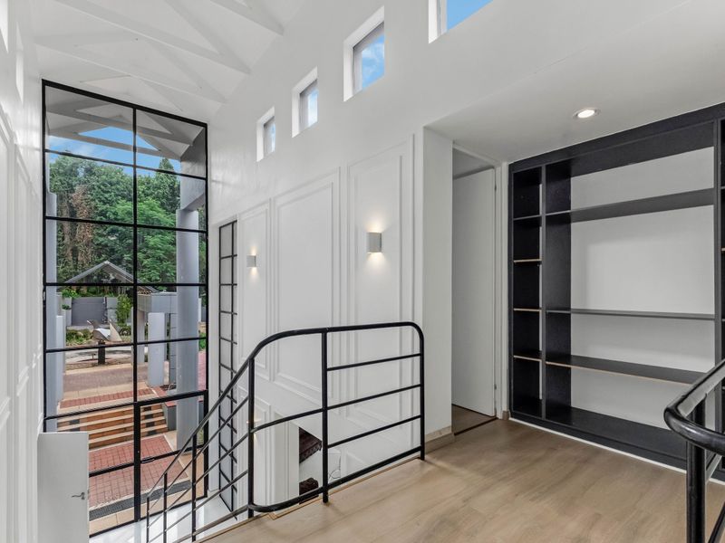 A bright, modern hallway featuring high vaulted ceilings, built-in shelving, and a large steel-framed window overlooking the outdoor area.