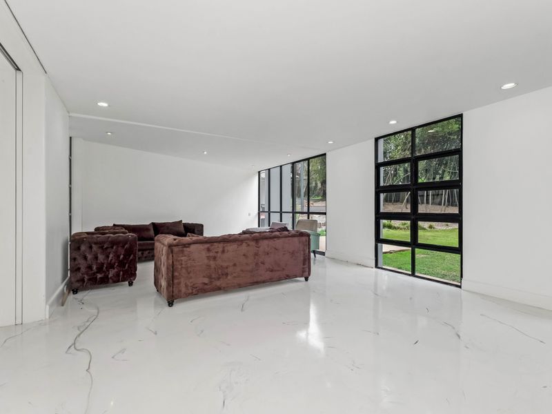 Spacious living area featuring polished marble-look flooring, recessed lighting, and large black-framed windows providing views of the garden.