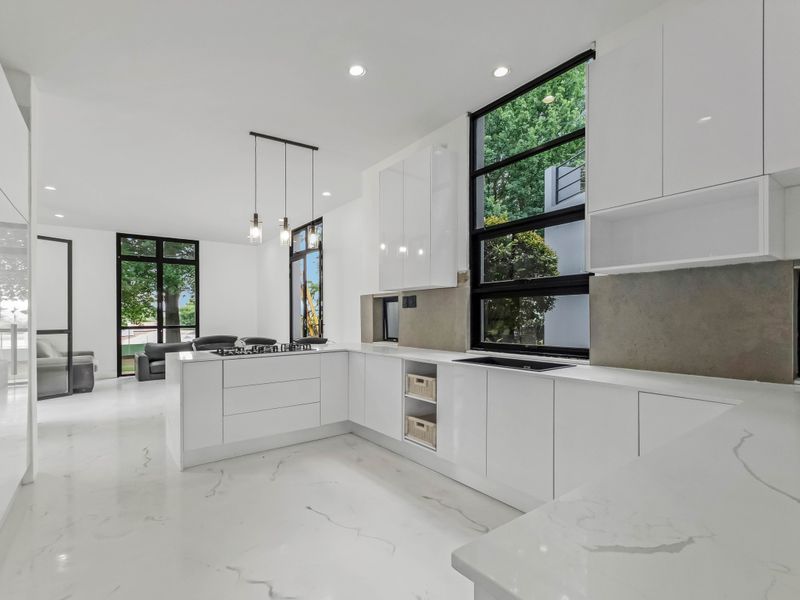 Modern open-plan kitchen featuring sleek white gloss cabinetry, stone countertops, integrated appliances, and large windows overlooking the garden.