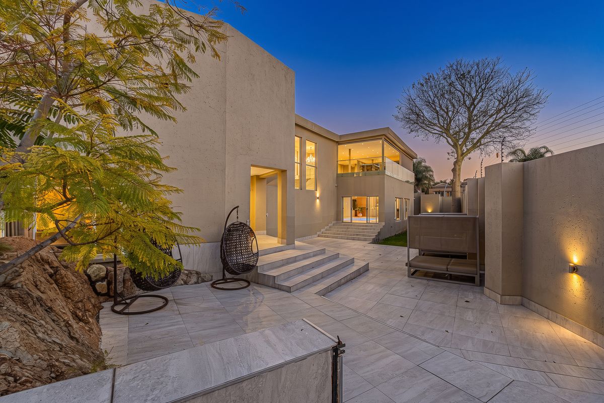 A modern rear exterior view featuring a spacious tiled patio, stone retaining wall, and elegant architectural lighting at dusk.