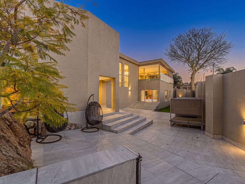 A modern rear exterior view featuring a spacious tiled patio, stone retaining wall, and elegant architectural lighting at dusk.