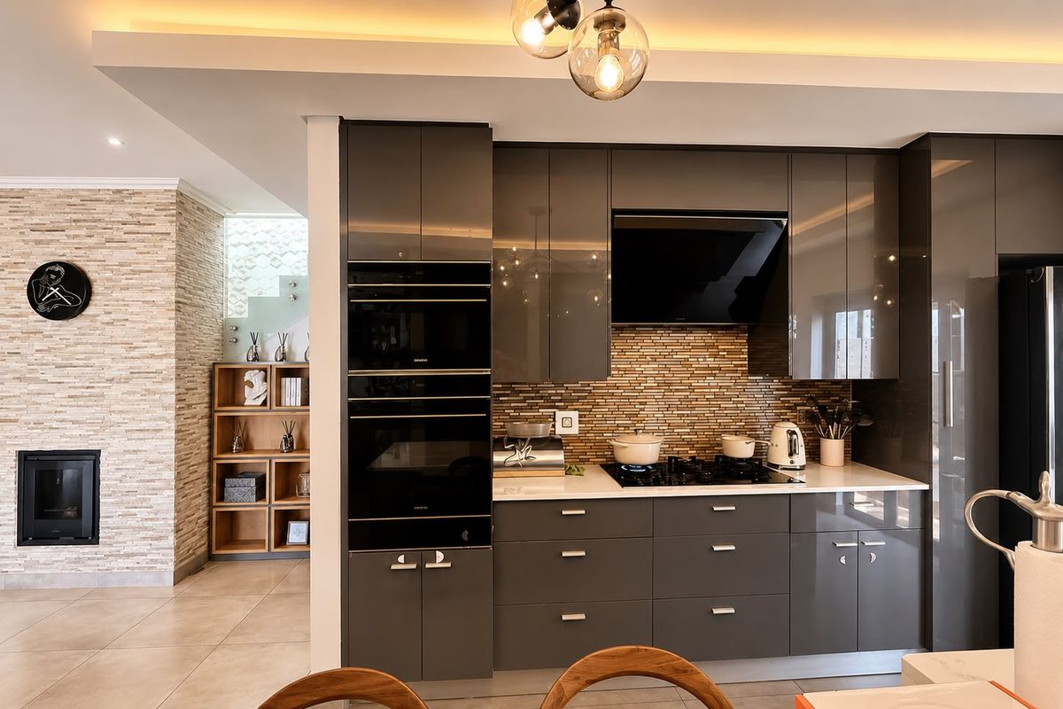 Modern kitchen featuring high-gloss grey cabinetry, integrated appliances, stone countertops, and a tiled backsplash, adjacent to a living area with a stone-clad fireplace.