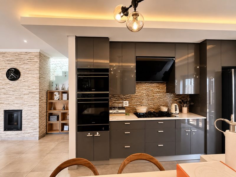 Modern kitchen featuring high-gloss grey cabinetry, integrated appliances, stone countertops, and a tiled backsplash, adjacent to a living area with a stone-clad fireplace.