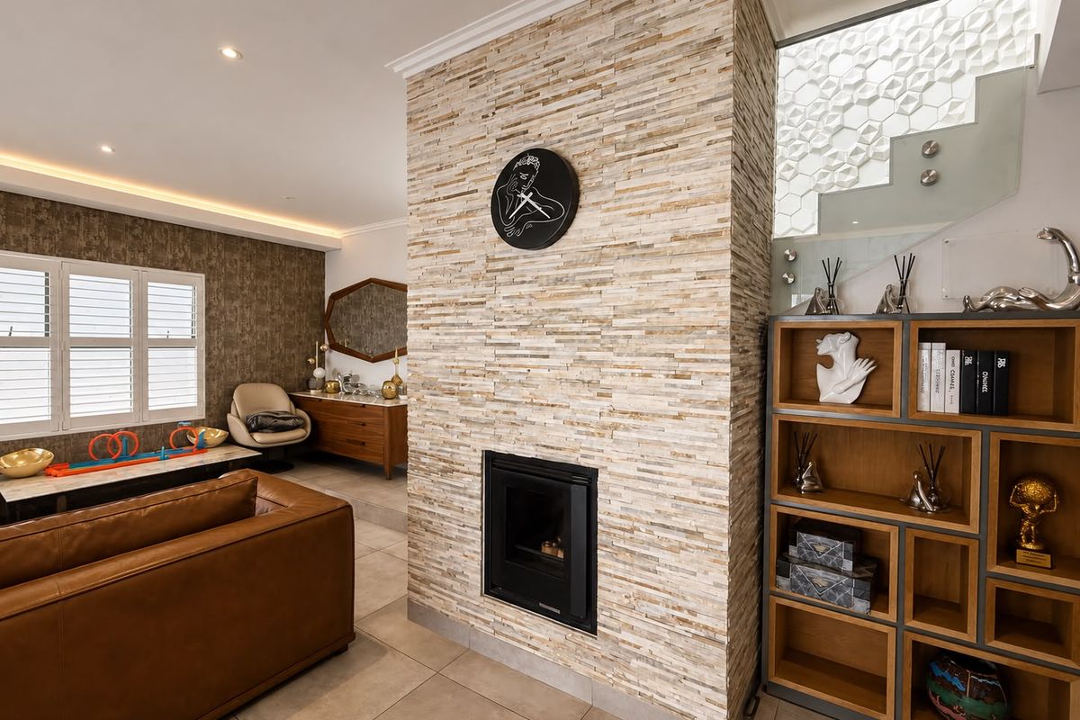 A modern living room featuring a prominent stone-clad fireplace, recessed lighting, and a stylish glass staircase with a textured feature wall.