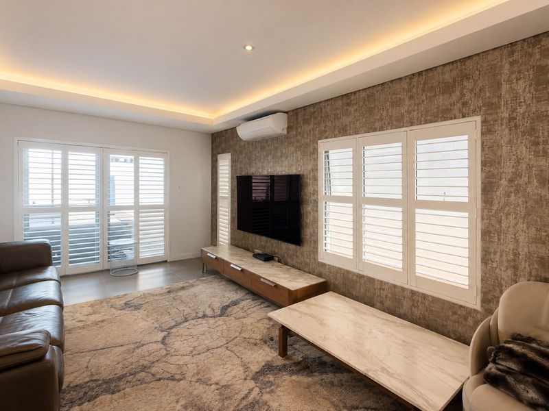A modern, well-maintained living room featuring elegant plantation shutters, recessed cove lighting, textured feature wallpaper, and sleek tiled flooring.