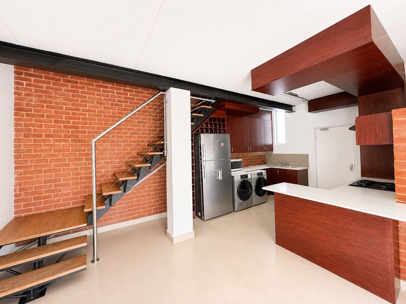Modern open-plan kitchen and living area featuring exposed brick walls, a contemporary staircase, and sleek wooden cabinetry with integrated appliances.