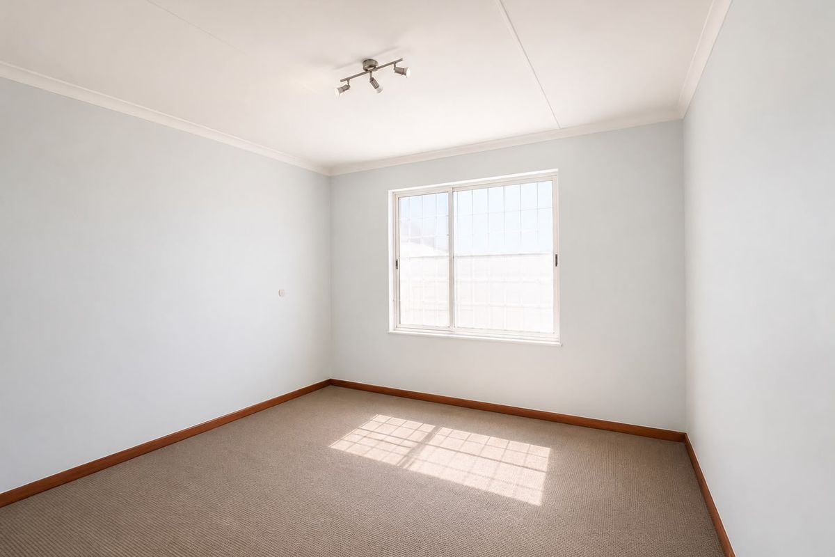 A clean, empty bedroom featuring neutral carpeting, a sliding window with security bars, and simple track lighting.