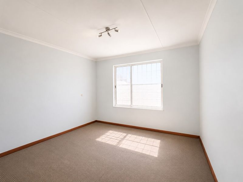 A clean, empty bedroom featuring neutral carpeting, a sliding window with security bars, and simple track lighting.