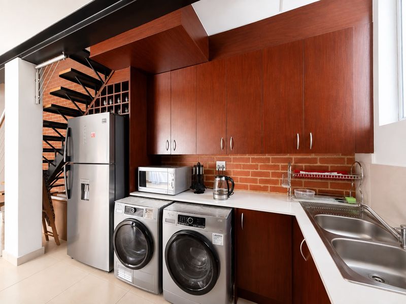 A functional laundry area featuring dark wood cabinetry, exposed brick accents, a stainless steel double sink, and integrated space for laundry appliances.