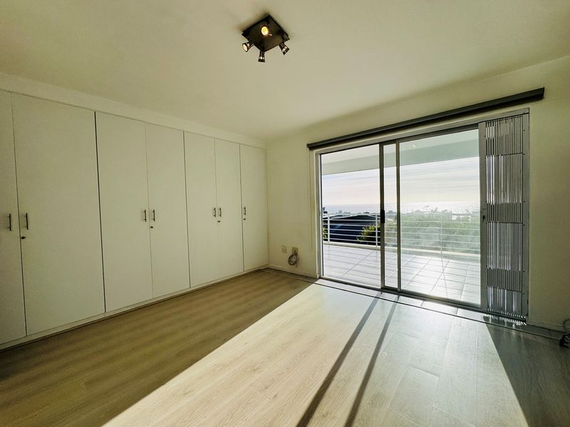 Bright bedroom featuring extensive built-in cupboards, light laminate flooring, and a sliding glass door leading to a balcony with ocean views.