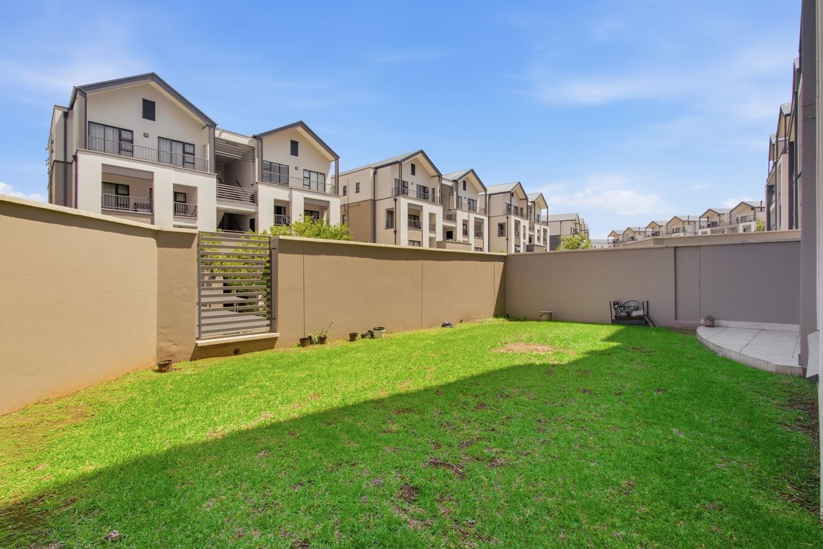 A private rear garden featuring a well-maintained lawn, boundary walls, and a small tiled patio area, set within a modern apartment complex.