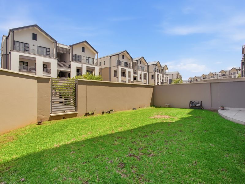 A private rear garden featuring a well-maintained lawn, boundary walls, and a small tiled patio area, set within a modern apartment complex.