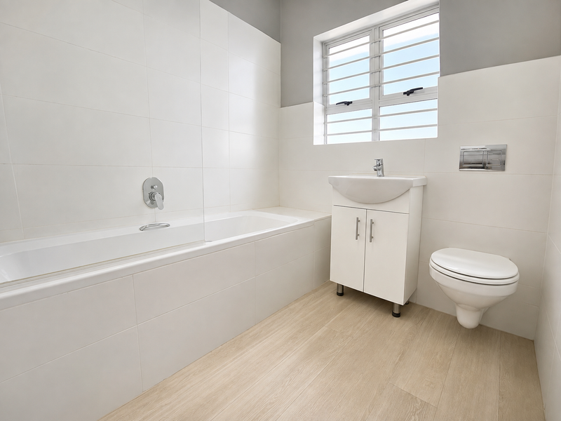 A modern, clean bathroom featuring a built-in bathtub, wall-mounted toilet, vanity basin, and neutral tiled finishes with wood-look flooring.