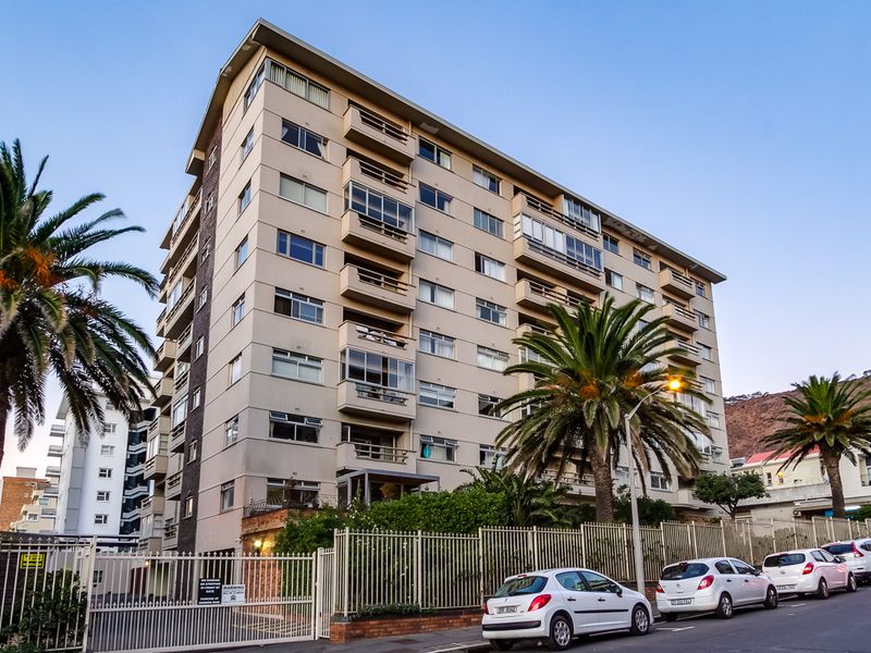 Exterior view of a multi-story apartment building featuring balconies and secure perimeter fencing, situated along a street.