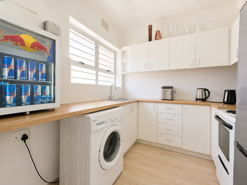 A functional kitchen featuring white cabinetry, laminate countertops, tiled splashbacks, and integrated laundry plumbing, complemented by a window with security bars.